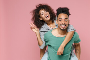 Fototapeta premium Funny young african american couple friends guy girl in gray green casual clothes giving piggyback ride to joyful, sitting on back showing victory sign isolated on pink background studio portrait.