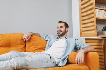 Smiling handsome attractive pleasant young bearded man 20s wearing casual white t-shirt blue shirt looking aside sitting on couch resting relaxing spending time in living room at home.