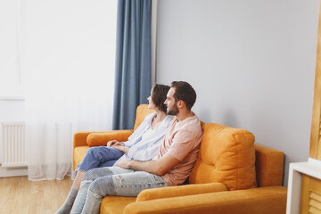 Side view of smiling attractive beautiful young couple two friends man woman 20s wearing casual clothes sitting on couch hugging looking aside resting relaxing spending time in living room at home.