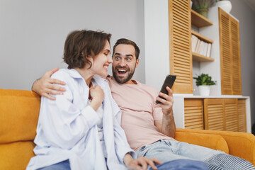 Excited surprised shocked cheerful young couple two friends man woman 20s wearing casual clothes sitting on couch hugging using mobile cell phone resting relaxing spending time in living room at home.