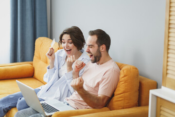 Overjoyed young couple two friends man woman in casual clothes sit on couch doing winner gesture using laptop pc computer hold credit bank card resting relaxing spending time in living room at home.