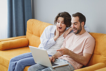 Shocked amazed excited young couple two friends man woman in casual clothes sitting on couch hugging pointing index finger on laptop pc computer resting relaxing spending time in living room at home.