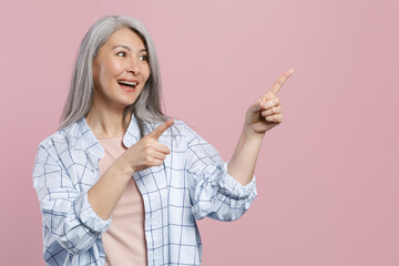 Cheerful funny gray-haired asian woman wearing basic white checkered shirt standing pointing index fingers aside up on mock up copy space isolated on pastel pink colour background, studio portrait.