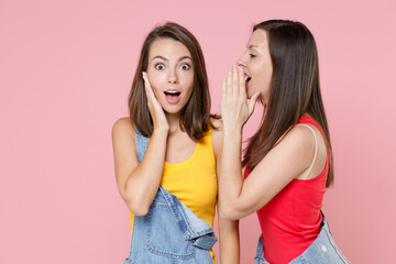 Two shocked amazed young women friends 20s in casual denim clothes posing whispering secret behind hand, sharing news put palm on cheek isolated on pastel pink colour background, studio portrait.