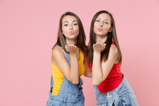 Two pretty charming attractive young women friends 20s wearing casual denim clothes posing standing hugging blowing sending air kiss isolated on pastel pink colour background, studio portrait.
