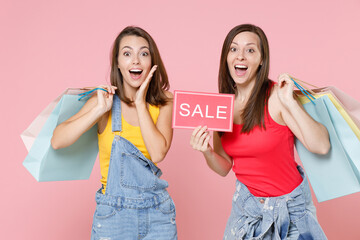 Two surprised young brunette women friends 20s in casual denim clothes hold package bag with purchases after shopping sign with SALE title isolated on pastel pink colour background studio portrait.