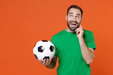 Pensive young man football fan in green t-shirt cheer up support favorite team with soccer ball put hand prop up on chin isolated on orange background studio. People sport leisure lifestyle concept.