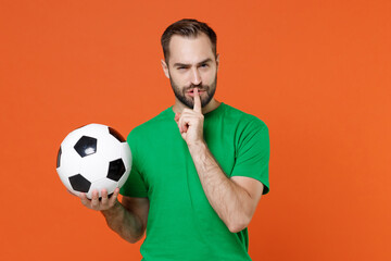 Secret man football fan in green t-shirt cheer up support favorite team with soccer ball say hush be quiet with finger on lips shhh gesture isolated on orange background. People sport leisure concept.