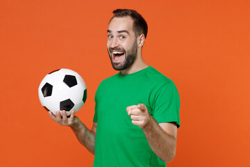 Cheerful young man football fan in green t-shirt cheer up support favorite team with soccer ball pointing index finger on camera isolated on orange background. People sport leisure lifestyle concept.