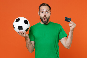 Shocked young man football fan in green t-shirt cheer up support favorite team with soccer ball hold credit bank card isolated on orange background in studio. People sport leisure lifestyle concept.