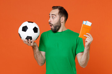 Amazed young man football fan in green t-shirt cheer up support favorite team with soccer ball hold passport ticket boarding pass isolated on orange background. People sport leisure lifestyle concept.