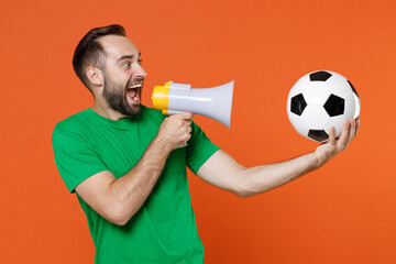 Excited young man football fan in green t-shirt cheer up support favorite team with soccer ball scream in megaphone looking aside isolated on orange background. People sport leisure lifestyle concept.