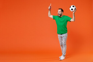 Fototapeta na wymiar Full length portrait screaming young man football fan in green t-shirt cheer up support favorite team with soccer ball hold beer bottle isolated on orange background. People sport leisure concept.