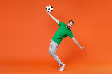 Fototapeta na wymiar Full length portrait funny man football fan in green t-shirt cheer up support favorite team with soccer ball spreading hands stand on toes isolated on orange background. People sport leisure concept.