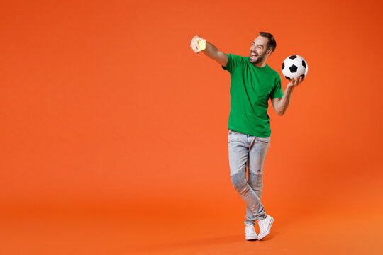 Full length portrait funny man football fan in basic green t-shirt support favorite team with soccer ball doing selfie shot on mobile phone isolated on orange background. People sport leisure concept.