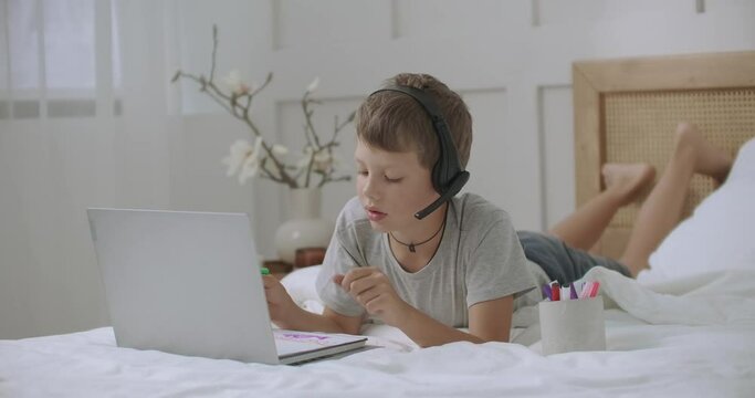 teen boy is coloring and drawing pictures at album lying on bed at home and communicating by laptop