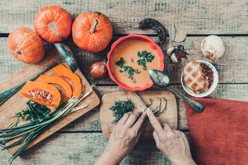 Pumpkin soup on wooden table with red linen cloth and vintage cutlery. Woman hands cutting vegetables, cooking process. Autumn vegetarian, healthy food concept. Thanksgiving dinner, top view.