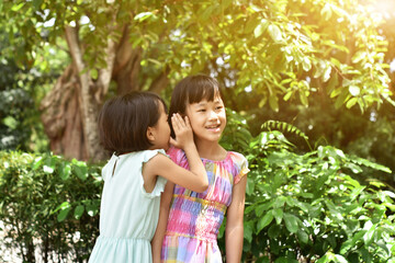 Two little sister girls whisper in ear at park outdoors. Friendship, happiness and people concept