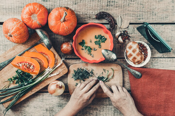 Pumpkin soup on wooden table, vintage. Woman hands, herbs, cooking process. Girl looking for a meal recipe on mobile phone while cooks at home. Thanksgiving, vegetarian, autumn. Flat lay, top view.