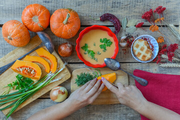 Pumpkin soup on wooden table with red linen cloth and vintage cutlery. Woman hands cutting vegetables, cooking process. Autumn vegetarian, healthy food concept. Thanksgiving dinner, top view.
