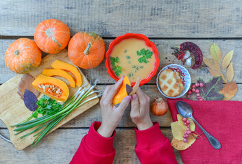 Pumpkin soup on wooden table with red linen cloth and vintage cutlery. Woman hands cutting vegetables, cooking process. Autumn vegetarian, healthy food concept. Thanksgiving dinner, top view.
