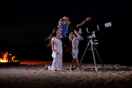 Happy family looking at moon and stars through a telescope at beautiful night sky. Parents and two children star gazing on the beach
