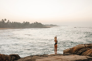 Back view young woman in black swimsuit and straw hat stand on rocky seashore and enjoy beautiful sea view