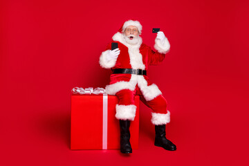 Full length body size view of his he handsome fat bearded amazed Santa sit on large big giftbox using device plastic card celebrate order shop isolated bright vivid shine vibrant red color background