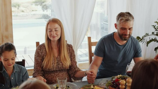 Tracking shot of mixed-aged family members sitting with eyes closed, holding hands and praying before eating holiday dinner at home