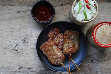 top view photo of juicy grilled pork or moo ping in Thai language in the black round dish on the wooden table with a sticky rice basket , chilli and sauce