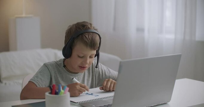 schooler boy is viewing learning video by notebook and listening teacher by headphones, drawing in copybook