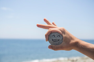 a hand holds a stone with a painted happy face, in the background you can see the sea
