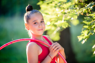 Portrait of a gymnast girl in a red jumpsuit with a hoop in the park against