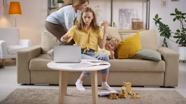 Handheld shot of woman sitting on couch and looking through documents. She is closing laptop in frustration and burying her head in her hands, then leaving while children playing and making noise
