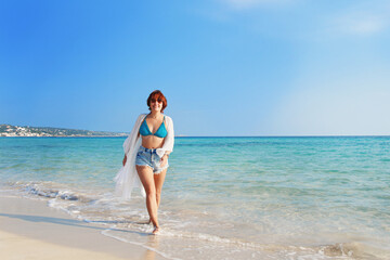 Fototapeta na wymiar Young woman in cover up and jeans shorts walking on the beach