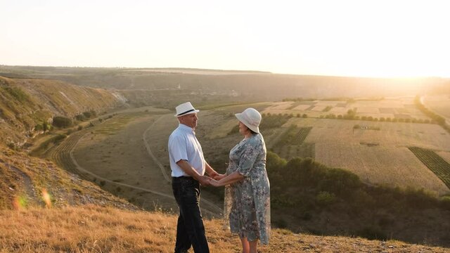 Senior couple stay on the top of the hill and admire the sunset while holding hands and kissing each other. Elderly Family picnic at sunset.