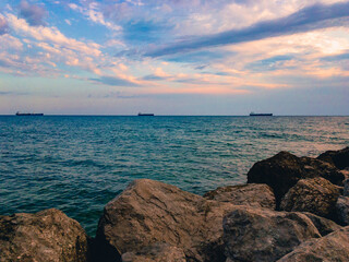 The surface of the sea against a blue cloudy sky with three ships on the horizon and large stones in the foreground.