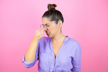 Young beautiful woman wearing glasses over isolated pink background smelling something stinky and disgusting, intolerable smell, holding breath with fingers on nose