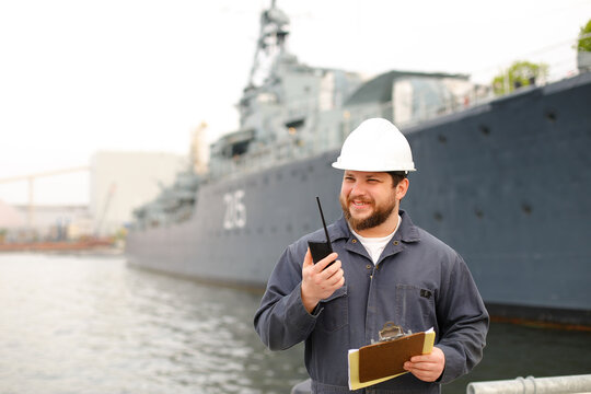 Marine first mate talking by walkie talkie and standing near vessel with documents.