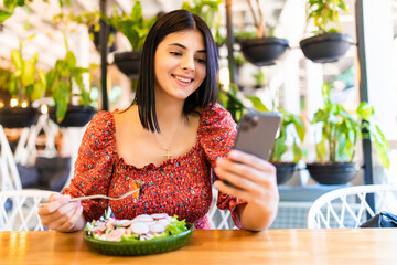 Beautiful young woman is using an application to send an sms message in her smartphone device while eating a salad at the restaurant