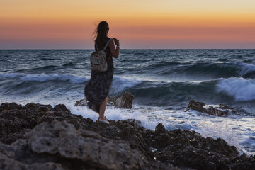 A girl with a backpack stands against the sea. A lone traveler admires the sunset. A dark-haired girl watches a storm at sea. Colorful atmospheric landscape.