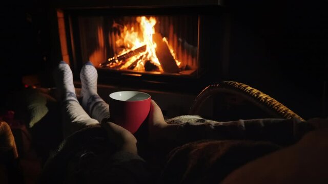 A man warms his feet by the fireplace, holds in his hands a red cup with a hot drink