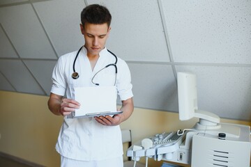 Charming young male doctor , sitting at his office near ultrasound scanning machine. Handsome friendly doctor enjoying working at the hospital, copy space. Ultrasonic concept