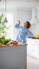 Vertical Shot of Energetic Young Man with Long Hair Dancing in the Kitchen while Wearing Blue Pajamas. Bright White Modern Kitchen Area with Healthy Green Vegetables on a Table. Cozy Home.