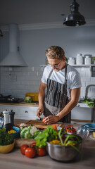 Handsome Man in White Shirt and Apron is Making a Healthy Organic Salad Meal in a Modern Sunny Kitchen. Hipster Man in Glasses Cooking. Natural Clean Diet and Healthy Way of Life Concept.
