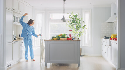 Energetic Funny Young Man with Long Hair Dancing in the Kitchen while Wearing Blue Pajamas. Bright White Modern Kitchen Area with Healthy Green Vegetables on a Table. Cozy Home.