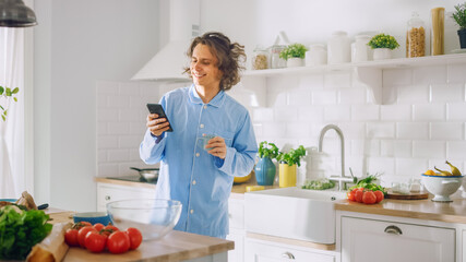 Happy Young Man with Long Hair is Using Smartphone in a Kitchen while Wearing Blue Pajamas. He is Scrolling Social Meadia and News Feed. Energetic Man Drinking Coffee and Having a Healthy Breakfast.