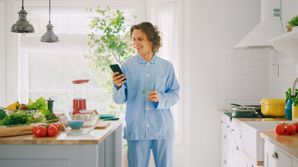 Happy Young Man with Long Hair is Using Smartphone at Home while Wearing Blue Pajamas. He is Holding Cup of Coffee. Energetic Man Scrolling News Feed and Checking Social Media Notifications.
