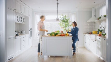 Two Young Handsome Men Talk in the Kitchen. One is Dressed Casually and His Friend Wears a Business Suit. Sunny Modern Kitchen with Healthy Green Vegetables on Table. Happy Gay Couple at Home.