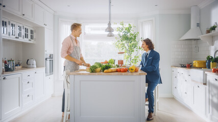 Two Young Handsome Men Talk in the Kitchen. One is Dressed Casually and His Friend Wears a Business Suit. Sunny Modern Kitchen with Healthy Green Vegetables on Table. Happy  Gay Couple at Home.